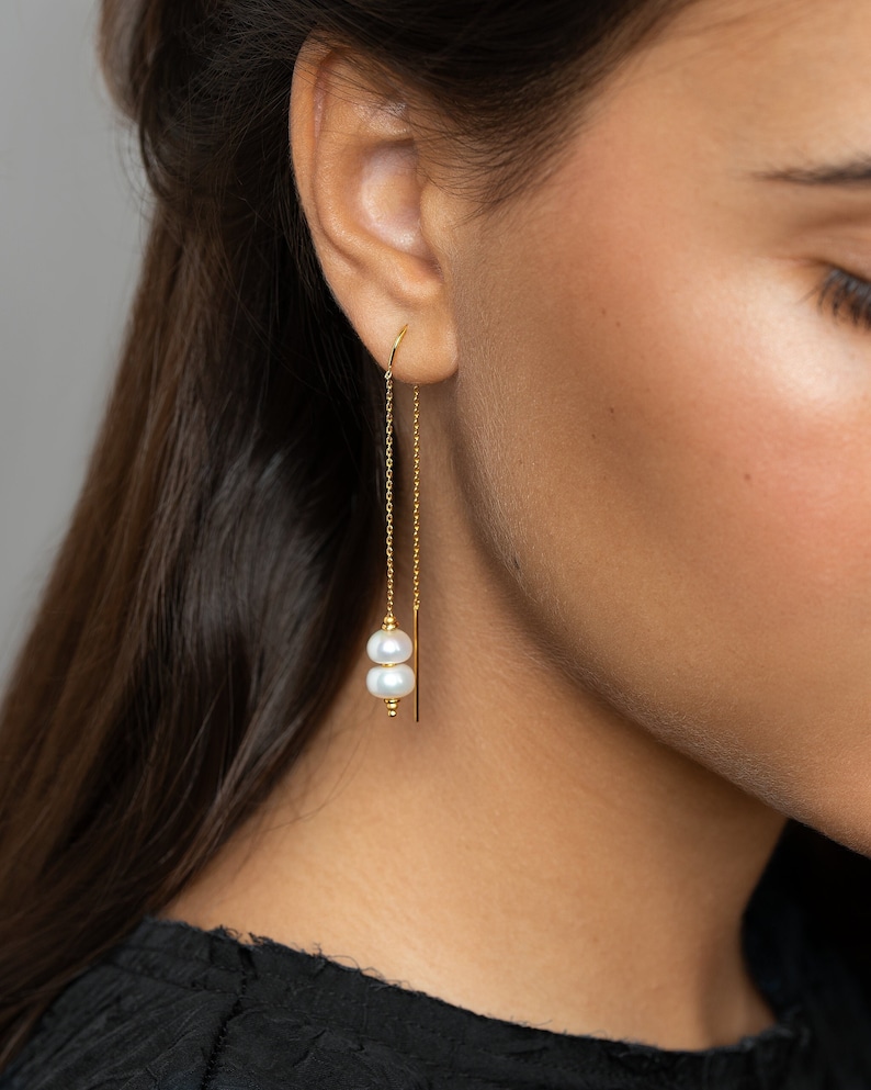 May include: Gold chain earrings with two white pearls hanging from the bottom. The earrings are worn by a person with long brown hair.