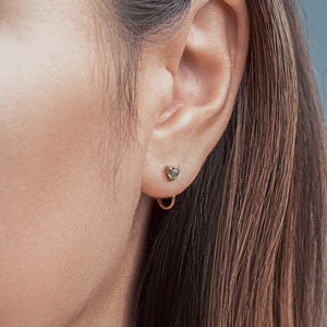 Puede incluir: Primer plano de una oreja con un pequeño pendiente de piedra gris oscuro y un aro de oro.