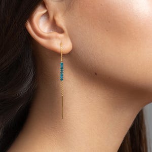 Pode incluir: Um brinco de fio dourado com uma corrente fina que passa pelo lóbulo da orelha. O brinco é adornado com quatro pequenas contas azuis facetadas e vibrantes ao longo da corrente, terminando numa barra de metal dourada fina e reta.