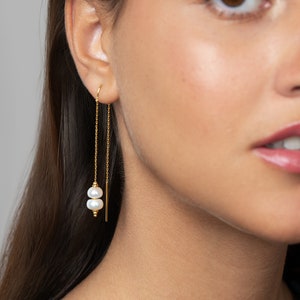 May include: Gold chain earrings with two white pearls hanging from the chain. The earrings are dangling from the earlobe.