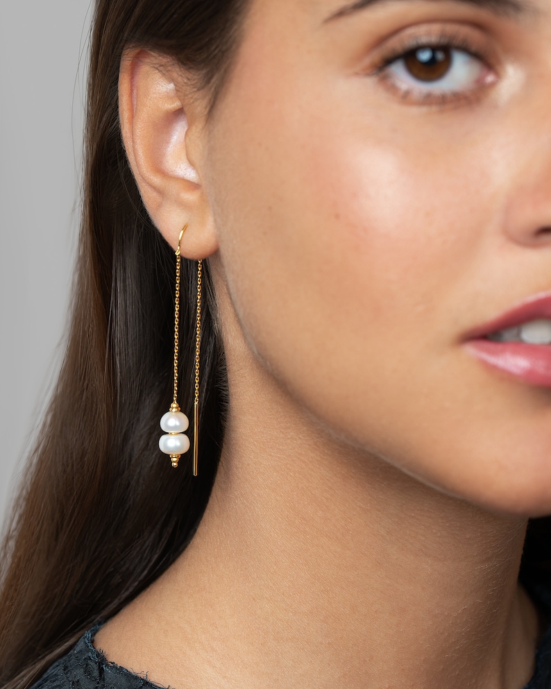 May include: Gold chain earrings with two white pearls hanging from the chain. The earrings are dangling from the earlobe.