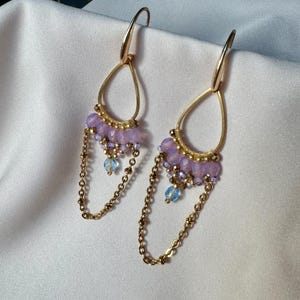 May include: A pair of gold-tone earrings with a teardrop shape. The earrings feature a cluster of small, light purple beads and dangling gold chains. Small blue beads are also incorporated into the design. The earrings are displayed on a white satin surface.