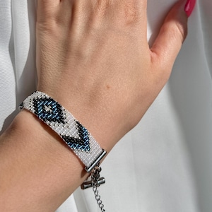 May include: White beaded bracelet with a blue and black eye design. The bracelet has a silver-colored clasp and chain. The bracelet is on a wrist.