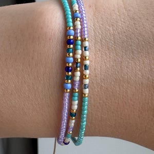 May include: Three layered bracelets made of small seed beads in shades of turquoise, lavender, white, and dark blue, accented with small metallic gold beads. The bracelets feature a thin cord closure with a dangling bead at the end of the string.