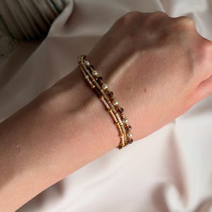 May include: A multi-strand beaded bracelet featuring a combination of small metallic gold beads, cream-coloured seed beads, white pearls, and translucent dark purple faceted beads arranged in a delicate, layered design on a thin cord.
