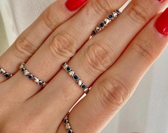 May include: Four silver-toned rings worn on fingers, each featuring a repeating pattern of small metallic hearts alternating with tiny, dark, circular stones. The bands are thin and circular, encircling the fingers with a consistent, delicate design.