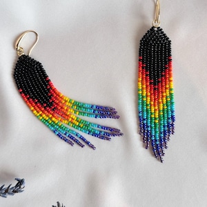 Op de afbeelding: Een paar kralen oorbellen met een regenboog franje ontwerp. Het bovenste deel van de oorbellen is zwart, overgaand in rood, oranje, geel, groen, blauw en paars. De oorbellen hebben goudkleurige haken.
