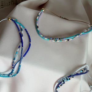 Op de afbeelding: Drie meerstrengs kralenkettingen met kleine glaskralen in tinten blauw, turquoise, wit en lavendel, geaccentueerd met kleine metallic gouden en zilveren tussenkralen. Elke ketting is voorzien van een kleine gouden of zilveren kraalsluiting.