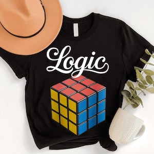 May include: Black t-shirt with the word "Logic" in white script above a colorful Rubik's Cube graphic. The cube features red, yellow, and blue squares. A tan hat and a white mug are in the background.