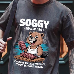 Könnte beinhalten: Dunkelgraues T-Shirt mit einem Cartoon-Biber, der eine Schürze trägt und Rippchen hält. Der Text "SOGGY BEAVER BBQ" steht über dem Biber, und "IF IT'S NOT ALL OVER YOUR FACE, YOU'RE EATING IT WRONG" steht darunter.