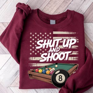 May include: Burgundy sweatshirt featuring a graphic of a pool table, the American flag, and the text "SHUT UP AND SHOOT." The design includes a black 8-ball and a rack of billiard balls. The sweatshirt is made of a soft material.