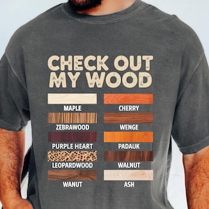 Puede incluir: Camiseta gris oscuro con el texto "CHECK OUT MY WOOD" en una fuente desgastada. Debajo, muestras de madera etiquetadas como Arce, Cerezo, Zebrano, Wengué, Corazón Púrpura, Padauk, Madera de Leopardo, Nogal, Wanut y Fresno.