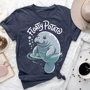May include: A navy blue t-shirt featuring a cartoon manatee design. The manatee is light gray and blue, with the words "Floaty Potato" in white script above it. The manatee is in water with bubbles around it. The shirt is folded.
