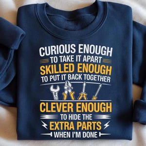 May include: Navy blue sweatshirt with white and yellow text that reads "CURIOUS ENOUGH TO TAKE IT APART, SKILLED ENOUGH TO PUT IT BACK TOGETHER, CLEVER ENOUGH TO HIDE THE EXTRA PARTS WHEN I'M DONE." The design includes illustrations of tools.