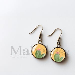 May include: A pair of round earrings with a cactus design. The earrings have a bronze-colored hook and a yellow background with a green cactus and pink flower. The earrings are a cute and unique accessory.