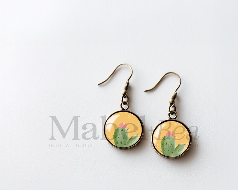 May include: A pair of round earrings with a cactus design. The earrings have a bronze-colored hook and a yellow background with a green cactus and pink flower. The earrings are a cute and unique accessory.