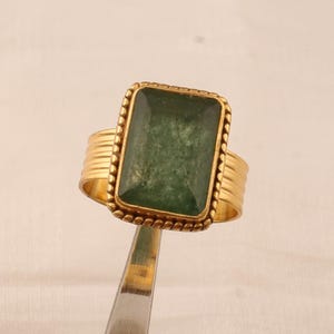May include: A gold-colored ring with a large, rectangular, green gemstone. The ring has a textured band and a beaded border around the stone. The ring is held by a silver tool against a white background.