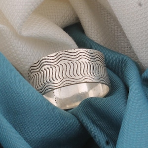 May include: A silver ring with a wavy pattern. The ring is displayed on teal and white fabric. The ring's design features a series of parallel, curved lines. The ring's design is a series of parallel, curved lines.