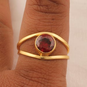 May include: A gold ring with a large, round, deep red gemstone. The ring's design includes a double band that curves around the stone, creating an eye-like shape. The ring is set on a finger.