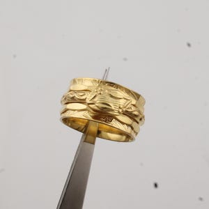 May include: A gold-colored ring with intricate floral and geometric patterns. The ring is held by a pair of tweezers, showcasing its detailed design. The ring appears to be made of metal and is a piece of jewelry.