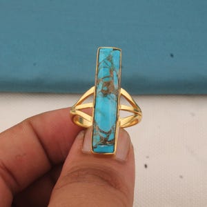May include: A gold-colored ring featuring a rectangular turquoise stone with brown veining. The ring has a split shank design. The turquoise stone is set in a gold bezel.