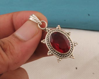 May include: A silver pendant with an oval, deep red gemstone. The pendant has a decorative border and a small loop at the top for a chain. The gemstone is the focal point, set in a detailed, ornate silver setting.