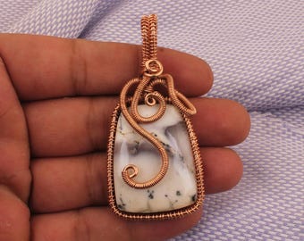 Peut inclure: Pendentif artisanal avec une pierre blanche rectangulaire parsemée de taches noires, sertie dans du fil de cuivre. Le fil est tissé de manière complexe autour de la pierre, formant un cadre décoratif et une bélière pour un collier. Le pendentif est tenu dans une main.