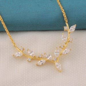 May include: A gold-colored necklace featuring a delicate leaf design. The necklace has a series of marquise-cut clear gemstones set along the leaf branches. The chain is a fine, gold-colored cable chain.