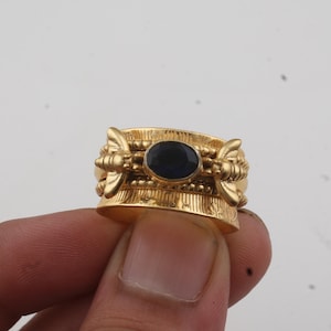 Op de afbeelding: Een goudkleurige ring met een ovale, donkerblauwe edelsteen in het midden. De ring heeft sierlijke details en wordt door een hand vastgehouden. Het ontwerp bevat decoratieve elementen aan de zijkanten.