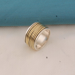 Puede incluir: Un anillo de plata con un diseño de banda dorada. El anillo presenta una banda ancha con múltiples rayas doradas paralelas, creando una apariencia texturizada. El anillo está sobre un fondo blanco y turquesa.