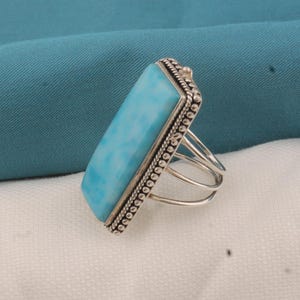 May include: A rectangular, light blue gemstone ring with a silver band. The gemstone is set in a silver bezel with a beaded border. The ring has a multi-band design.