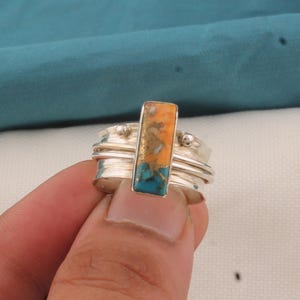 May include: A silver ring with a rectangular stone in shades of orange, gold, and turquoise. The ring has a spinning band design. The stone is the focal point, showcasing a unique colour combination. The ring is held by a person's hand.