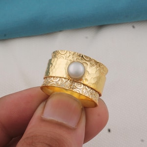 May include: A gold ring featuring a central pearl and a textured, layered design. The ring's bands have a hammered effect and detailed patterns. The pearl is a pale, creamy hue.