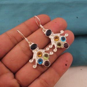 Puede incluir: Un par de pendientes de plata con un diseño único. Cada pendiente presenta una piedra ovalada negra en la parte superior, con un grupo de piedras más pequeñas en amarillo, verde, azul y burdeos. Los pendientes se sostienen en una mano.