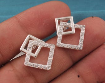 May include: A pair of silver stud earrings, each featuring two overlapping square frames. The outer frame is encrusted with small, clear gemstones. The earrings are displayed against a neutral background.