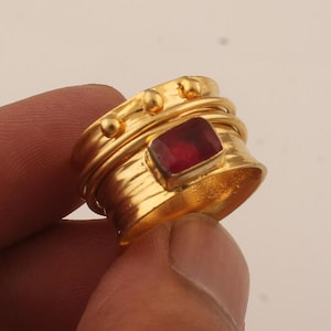 Op de afbeelding: Een goudkleurige ring met een rechthoekige, rode edelsteen in het midden. De ring heeft een getextureerde band en is versierd met kleine, ronde versieringen. De ring wordt door een persoon vastgehouden.