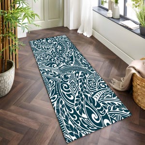 Puede incluir: Una alfombra rectangular con un patrón tribal turquesa y blanco. El diseño presenta líneas onduladas, formas de hojas y elementos geométricos. La alfombra está colocada sobre un suelo de madera oscura.