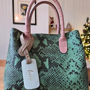 Peut inclure: Sac à main vert et noir à motif peau de serpent, avec des poignées et des détails roses. Le sac comporte une étiquette bleu clair avec le nom de la marque. Les poignées sont de couleur rose clair. Le sac est posé sur une surface en bois.
