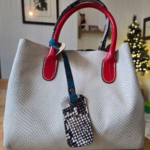 Puede incluir: Un bolso gris claro con un estampado de piel de serpiente. El bolso tiene asas rojas y una correa azul. Una etiqueta a juego con un estampado de piel de serpiente en blanco y negro y la letra "T" cuelga del asa. El bolso está sobre una superficie de madera.