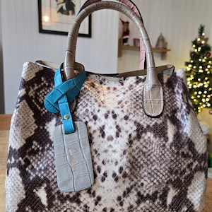 Peut inclure: Un sac à main à motif peau de serpent avec une étiquette et des poignées bleu clair. Le sac présente un design structuré avec une poignée supérieure et une étiquette détachable. La palette de couleurs comprend des nuances de marron, de blanc et de gris.