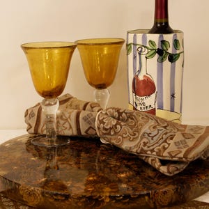 May include: A still life arrangement featuring two amber-colored wine glasses, patterned napkins, and a wine bottle in a decorative holder. The glasses sit on a round, brown, textured surface.