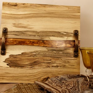 May include: A rectangular wooden serving tray with a natural wood grain pattern, accented by a strip of brown wood and bronze handles. Two amber-colored wine glasses are visible, suggesting a dining or entertaining theme.