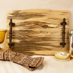 May include: A wooden serving tray with dark metal handles, two amber-colored wine glasses, a patterned napkin, a small bowl, and a wine bottle in a decorative cooler. The tray has a natural wood grain.