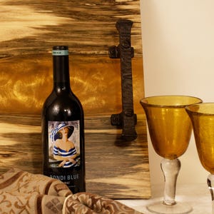 May include: A bottle of Bondi Blue Shiraz wine with a label featuring a woman in a hat and striped top. Two amber-colored wine glasses and a decorative wooden serving board are also visible.