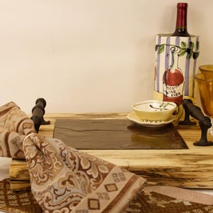 May include: A wooden serving tray with a slate insert, adorned with a wine bottle, two amber wine glasses, and a small teacup. Decorative napkins with a brown and beige pattern are draped around the tray. The wine bottle has a decorative sleeve.