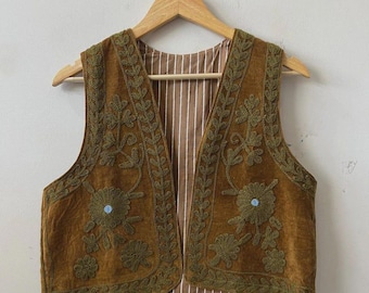 May include: A brown velvet vest with olive green embroidered floral designs and a striped interior. The vest is sleeveless and hangs on a wooden hanger. The floral embroidery includes small blue accents.