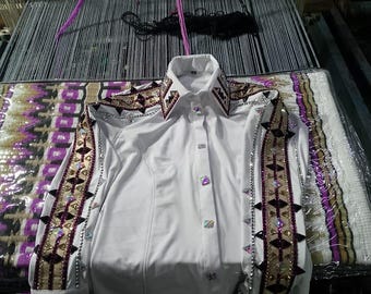May include: A white button-down shirt with ornate embellishments. The shirt features decorative patterns in gold, brown, and purple on the collar and sleeves. The sleeves are adorned with silver studs and rhinestones. The shirt is displayed on a loom.
