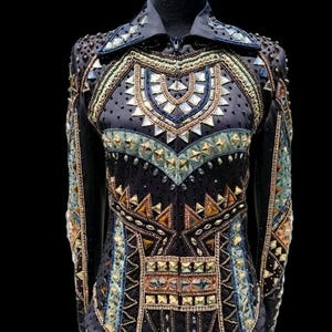 K&ouml;nnte beinhalten: Ein schwarzes Langarmshirt mit hohem Kragen und Frontrei&szlig;verschluss. Das Kleidungsst&uuml;ck ist reich verziert mit geometrischen Mustern aus Metallnieten, klaren Kristallen und bernsteinfarbenen Steinen, die in symmetrischen Reihen angeordnet sind.
