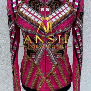 May include: A bright pink long-sleeve jacket featuring a high collar and a full-length front zipper. The garment is heavily embellished with geometric patterns of clear square crystals, black beadwork, and gold metallic accents. Text reads AJ ANSH INTERNATIONAL.