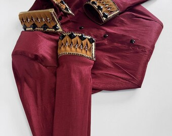 May include: A burgundy-colored garment with decorative gold and black embellishments on the cuffs and collar. The fabric has a subtle sheen. The garment appears to be a formal shirt or jacket, possibly for a special occasion.